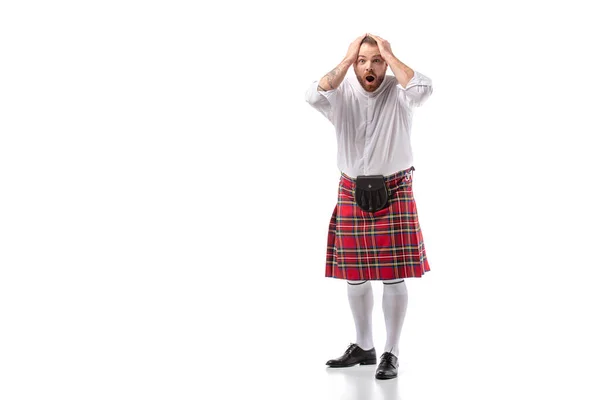 Homme barbu rousse écossaise choqué en kilt de tartan rouge tenant la tête avec la bouche ouverte sur fond blanc — Photo de stock