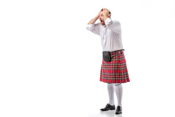 Шокированный шотландский рыжий бородатый мужчина в красном тартановом килте держа голову с открытым ртом на белом фоне — стоковое фото