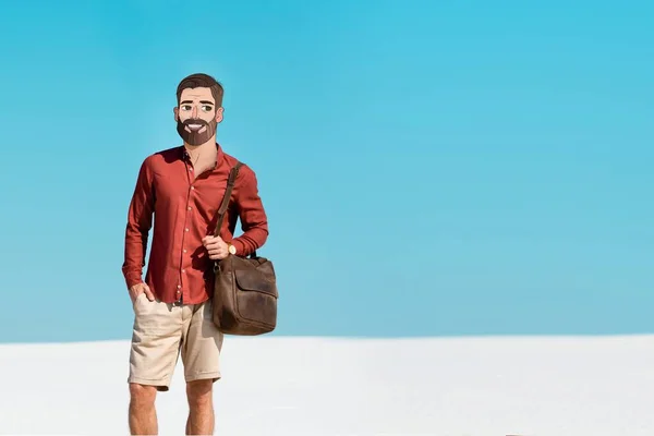 Homme avec visage illustré souriant tenant sac en cuir sur la plage de sable contre ciel bleu clair — Photo de stock