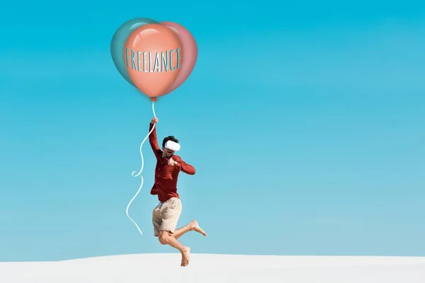 Uomo sulla spiaggia sabbiosa in auricolare vr che vola su palloncino con scritte freelance contro il cielo blu chiaro — Foto stock