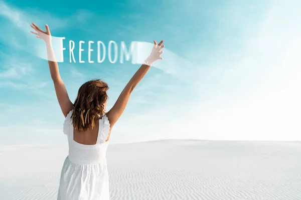 Back view of beautiful girl in white dress with hands in air on sandy beach with blue sky, freedom illustration — Stock Photo