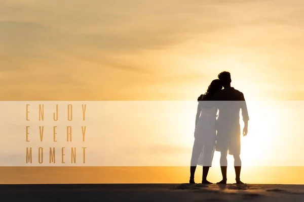 Silhuetas de homem e mulher abraçando na praia contra o sol durante o pôr do sol, aproveite cada momento ilustração — Fotografia de Stock