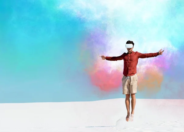 Man on sandy beach in vr headset jumping against clear blue sky, colorful clouds illustration — Stock Photo