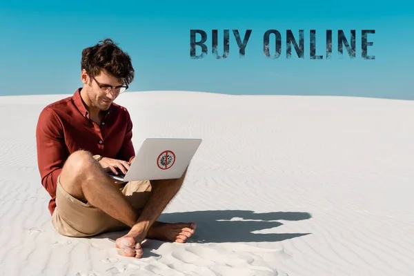 Молодой человек на песчаном пляже с помощью ноутбука против чистого голубого неба, купить онлайн иллюстрации — стоковое фото