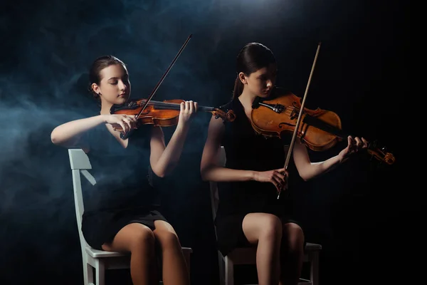 Attractive musicians playing on violins on dark stage with smoke — Stock Photo