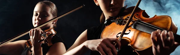 Attractive musicians playing on violins on dark stage with smoke, website header — Stock Photo
