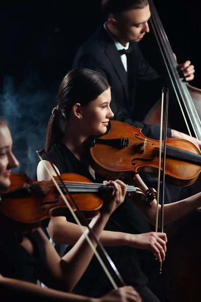 Trio of professional musicians playing on violins and contrabass on dark stage with smoke — Stock Photo