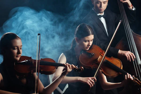 Трио музыкантов, играющих на скрипках и контрабасе на темной сцене с дымом — стоковое фото