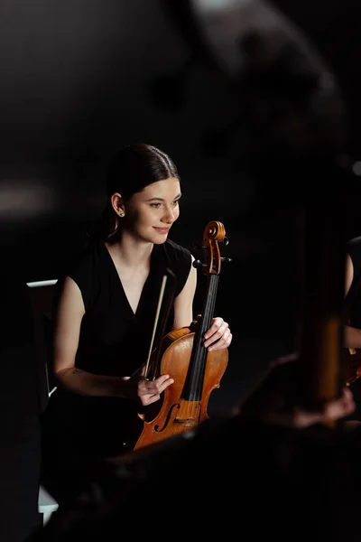 Feliz músico femenino sosteniendo el violín en el escenario oscuro - foto de stock