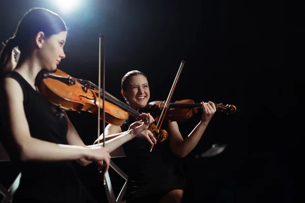 Laughing female musicians playing on violins on dark stage with back light — Stock Photo
