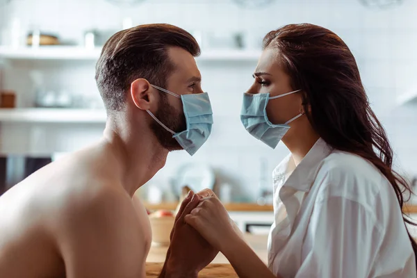 Side view of young couple in medical masks holding hands in kitchen — Stock Photo