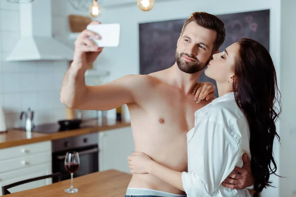 Женщина целует сексуального парня без рубашки, в то время как мужчина делает селфи на смартфоне на кухне — стоковое фото