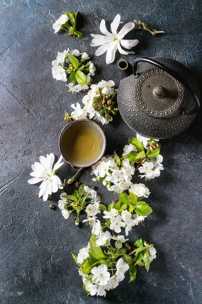 传统陶瓷杯的热绿茶与黑铁茶壶 春花白玉兰和樱桃盛开的树枝在深蓝色的纹理背景下 顶部视图 复制空间 — 图库照片