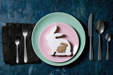 Paskalya sofrası, çatal bıçak takımı, boş tabak, tahta Paskalya dekorasyonu beyaz tavşan ve altın bıldırcın yumurtası. Klasik mavi doku arka planı. Düz uzanış, boşluk