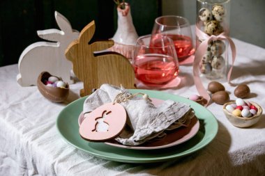 Tahta tavşan desenli Paskalya masası, çikolatalı yumurta ve tatlılar, pembe kokteyl bardakları, beyaz masa örtüsü üzerine boş tabaklar. Işıklı çelenkler. Tatil yemeği..