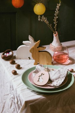 Tahta tavşan desenli Paskalya masası, çikolatalı yumurta ve tatlılar, pembe kokteyl bardakları, beyaz masa örtüsü üzerine boş tabaklar. Işıklı çelenkler. Tatil yemeği..