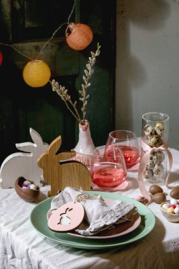 Tahta tavşan desenli Paskalya masası, çikolatalı yumurta ve tatlılar, pembe kokteyl bardakları, beyaz masa örtüsü üzerine boş tabaklar. Işıklı çelenkler. Tatil yemeği..