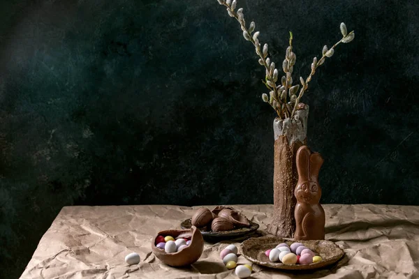 陶器の花瓶の花柳の枝 伝統的なチョコレートのウサギ 卵やお菓子とテーブルの上でイースター気分はまだ生活紙を砕いた 暗い背景 — ストック写真