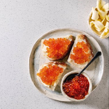 Kırmızı somon havyarı kasede ve buğday ekmeğinde, gri benekli arka planda seramik tabakta tereyağlı servis ediliyor. Düz uzanış, boşluk