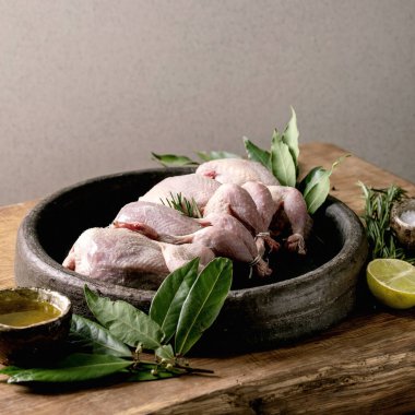 Raw uncooked quails ready for marinat in clay tray with lime, salt, pepper and greens over wooden cutting board.