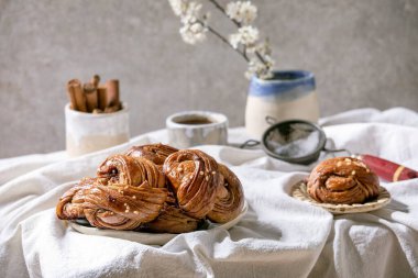 Geleneksel İsveç tarçınlı çörekler seramik tabakta Kanelbulle, bir sürahi şurup, beyaz katlanmış pamuk masa örtüsünde çiçek dalları. Boşluğu kopyala