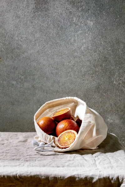 血淋淋的橙子 成熟多汁 装在棉花生态友好袋中 整片切碎 铺在灰色亚麻布桌布上 以混凝土墙为背景 — 图库照片