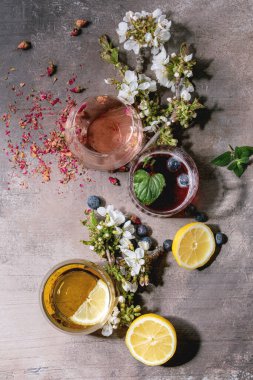 Parlak renkli içkilerle farklı kokteyl bardakları pembe gül şampanyası, elma şarabı veya limonata, limon, yabanmersini, nane. Yukarıda çiçek dalları var. Gri doku arka planı. Düz yatıyordu