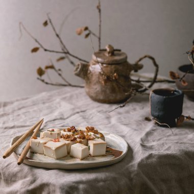 Japon usulü ipek tofu soya peyniri dilimlenmiş küp kırmızı biber, soğan ve soya sosu seramik tabakta yemek çubukları, seramik çaydanlık ve gri keten masa örtüsünde bardaklar. Asya yemeği.
