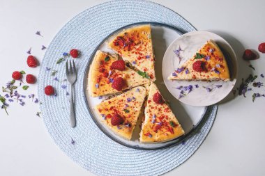 Dilimlenmiş ahududulu kek tabakta taze ahududu, yenilebilir çiçekler ve beyaz arka planda yuvarlak peçetede tatlı çatalıyla süslenmiş nane şekeri. Düz uzanış, boşluk