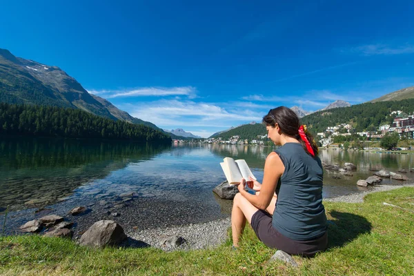 Mujer joven lee un libro frente a un lago de montaña — Foto de Stock