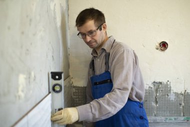 Tulumlu bir adam duvardaki gri çimentoya seramik fayanslar döşemek için mutfaktaki duvarları yıkıyor..