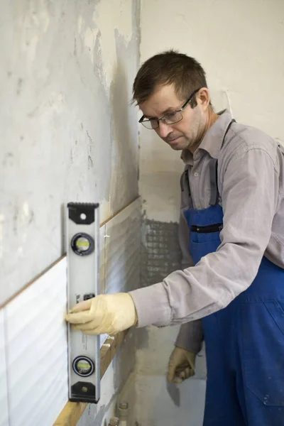 A man in overalls is leveling the walls in the kitchen to lay ceramic tiles on the gray cement on the wall. — Stock Photo, Image