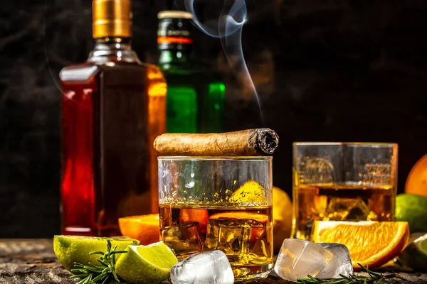 Alcohol de mano. concepto de alcoholismo, soledad, dependencia. Una botella de whisky, un vaso de whisky y con hielo, un cigarro con humo en una mesa negra. Bebida de élite para la relajación masculina — Foto de Stock