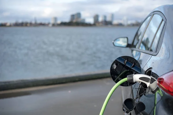 11-20-2020, Kopenhaga, Dania - Ładowanie samochodu elektrycznego, z miastem i wodą w tle, zielony kabel ładowania i czarny samochód. kopulacja — Zdjęcie stockowe