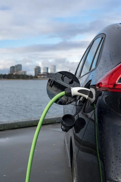 11-20-2020, Kopenhagen, Denemarken - Een elektrische auto opladen, met stad en water op de achtergrond, groene oplaadkabel en zwarte auto. zie Stockafbeelding
