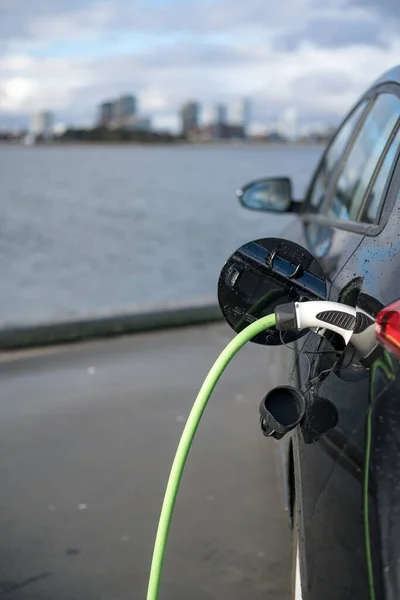 11-20-2020, Kodaň, Dánsko - Nabíjení elektromobilu s městem a vodou v pozadí, zeleným nabíjecím kabelem a černým automobilem. Kodaň Royalty Free Stock Obrázky