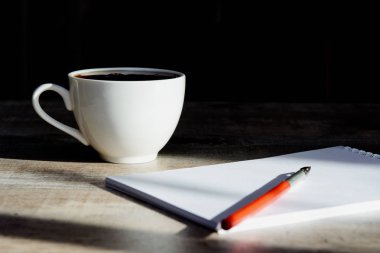 Masanın üzerinde bir defter, eski bir dolmakalem ve siyah bir fonda güneş tarafından aydınlatılmış bir fincan kahve var. Kavram - kahve molası, iş, muhasebeci