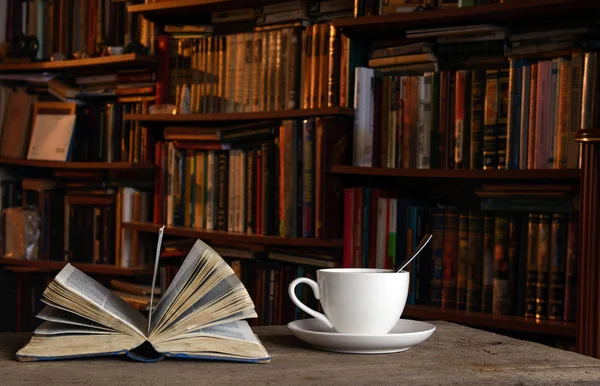 Açık kitap. Yakınlarda çay kaşığı ve çay tabağının üzerinde bir fincan kahve var. Arka planda kitaplarla birlikte. Kavram - kahve molası, eğitim, akşam okumaları