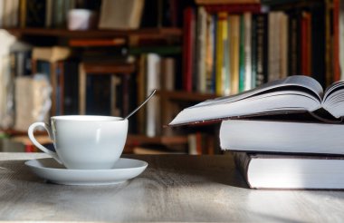 A white cup with coffee stands on a table lit by the sun. Opposite shelves with books. Nearby is an open book . Concept - writing, coffee break, winter evening