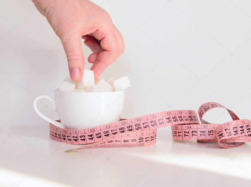 A coffee cup filled with lump sugar. In the female fingers a piece of sugar. Near a flexible meter. Sunlight. Concept - healthy eating, fitness, fighting obesity.