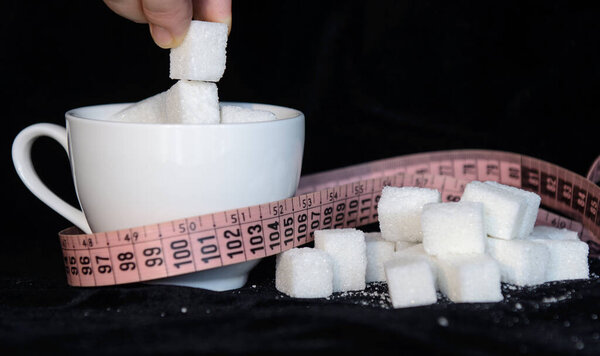 A white coffee cup filled with pieces of sugar. Around a measuring tape and pieces of sugar. Concept - monitor weight, limit sugar intake, diabetes, obesity