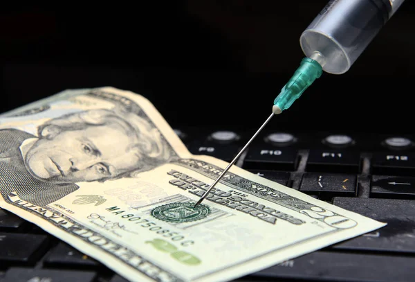On a computer keyboard is a twenty dollar bill. A syringe is inserted into the bill. Horizontal shot. The concept is the fall of the economy. Inflation, coronavirus pandemic. The collapse of stock indices.