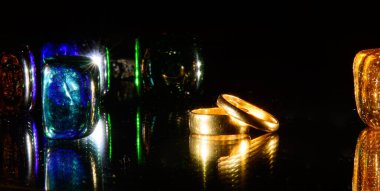 Siyah bir zemin üzerinde, renkli kristaller - yeşil, mavi, turuncu parlak ışıkla aydınlatılmış. Ayna. Neon ışığı. - Evet. Kristallerin arka planında, altın alyanslar. Konsept - Düğün ayini, nişan