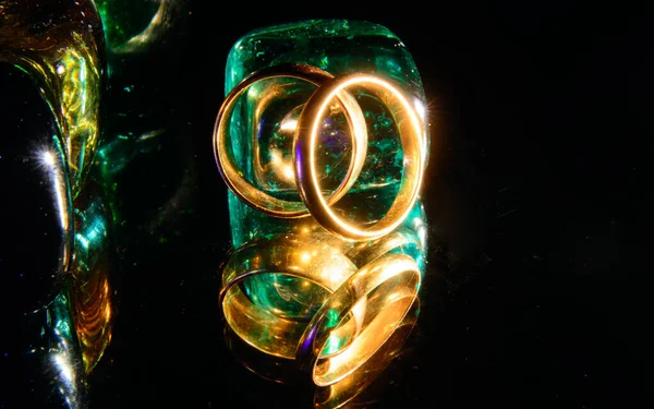 On a black background, colored crystals - green, blue, orange backlit with bright light. Mirror. Neon light. . On a background of crystals, gold wedding rings. Concept - sacrament of wedding, engagement