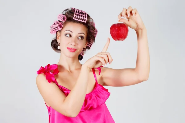 Huisvrouw met krulspelden in een rode nachtjapon en een appel in zijn hand. Grijze achtergrond — Stockfoto