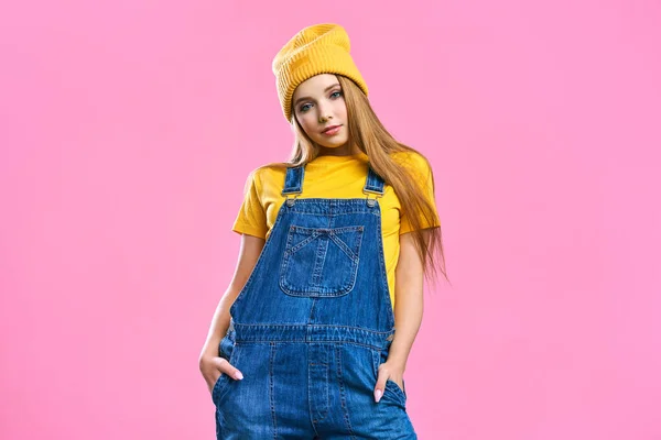 Portret en söt flicka i denim overaller och en gul hatt på en rosa bakgrund. Fashionistas kvinnliga student ler. Ljusa trendiga studio mode bild av sexig modell, bär neon ljusa färg block — Stockfoto