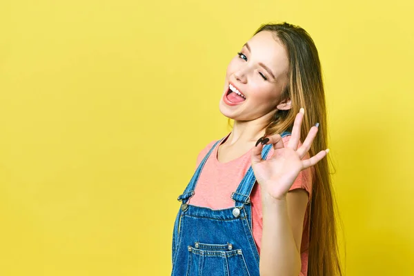 Vtipné a radostné dívka ukazuje v pořádku znamení a mrkne na kameru izolované na žlutém pozadí. — Stock fotografie