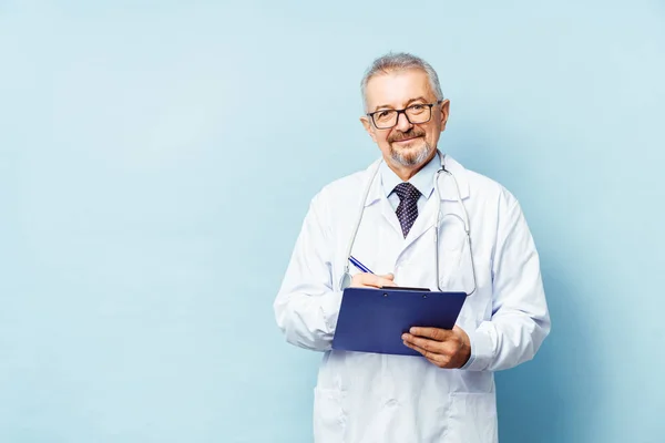 Médico mayor sonriente con estetoscopio. Sobre un fondo azul. El médico tiene la carpeta en sus manos y hace una cita en la clínica. Concepto de prevención —  Fotos de Stock