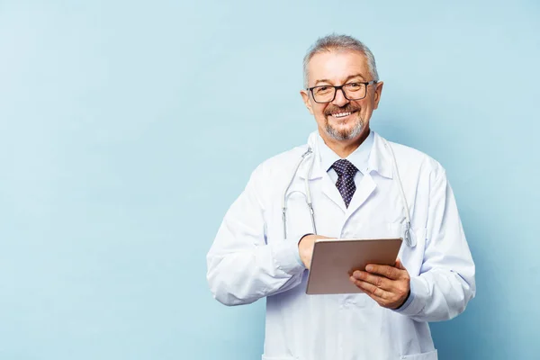 Médico senior con estetoscopio. Sobre un fondo azul. Un médico tiene una tableta en sus manos y hace una cita con la clínica. Concepto de prevención —  Fotos de Stock