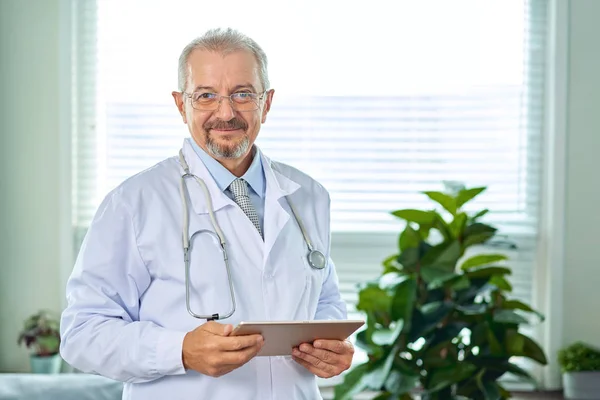Medical senior doctor with a stethoscope. On a blue background. A medic holds a tablet in his hands and makes an appointment with the clinic. Disease prevention concept